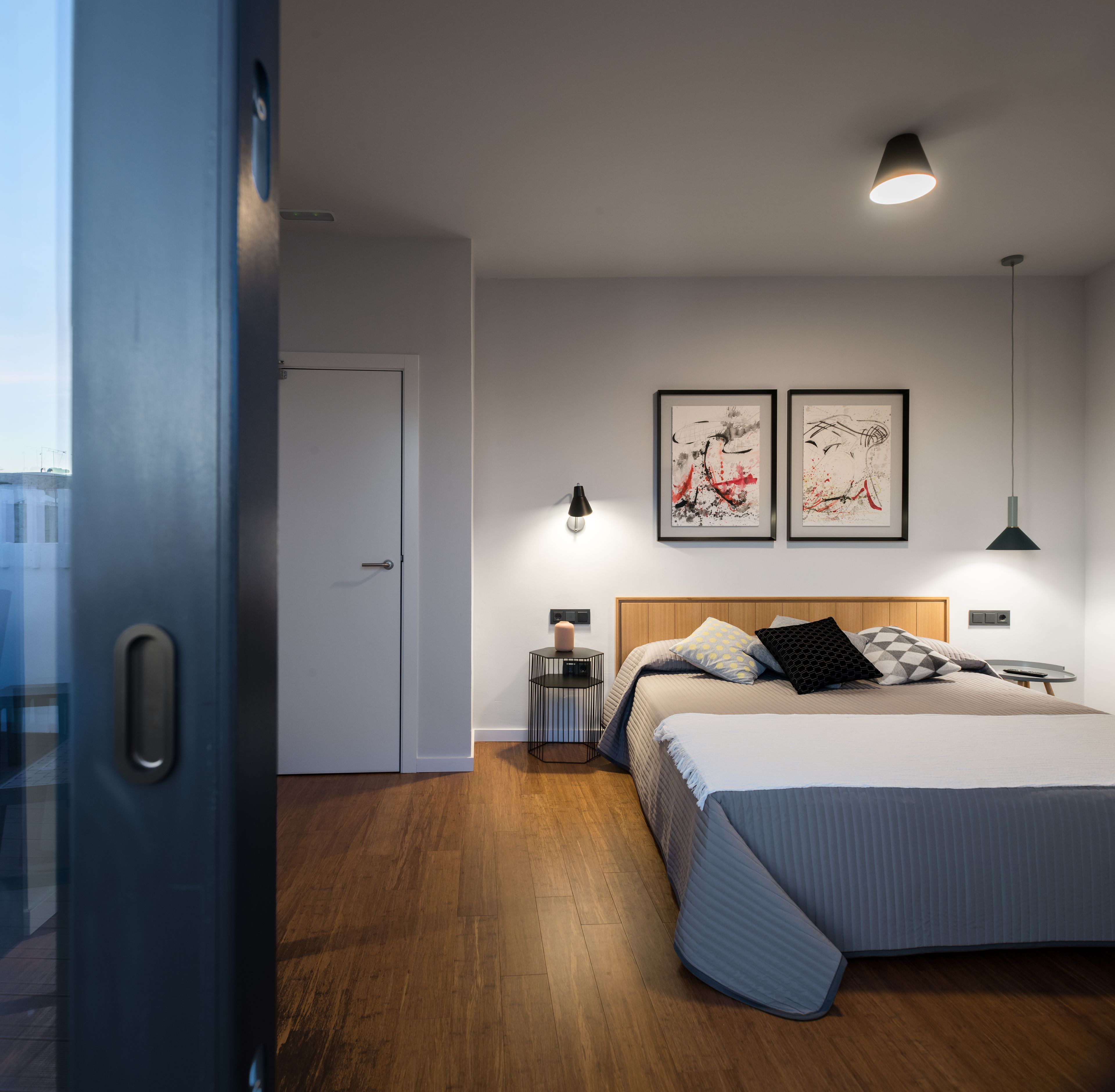 A modern bedroom with a large bed, wooden headboard, two framed artworks, bedside lamps, and a nightstand.