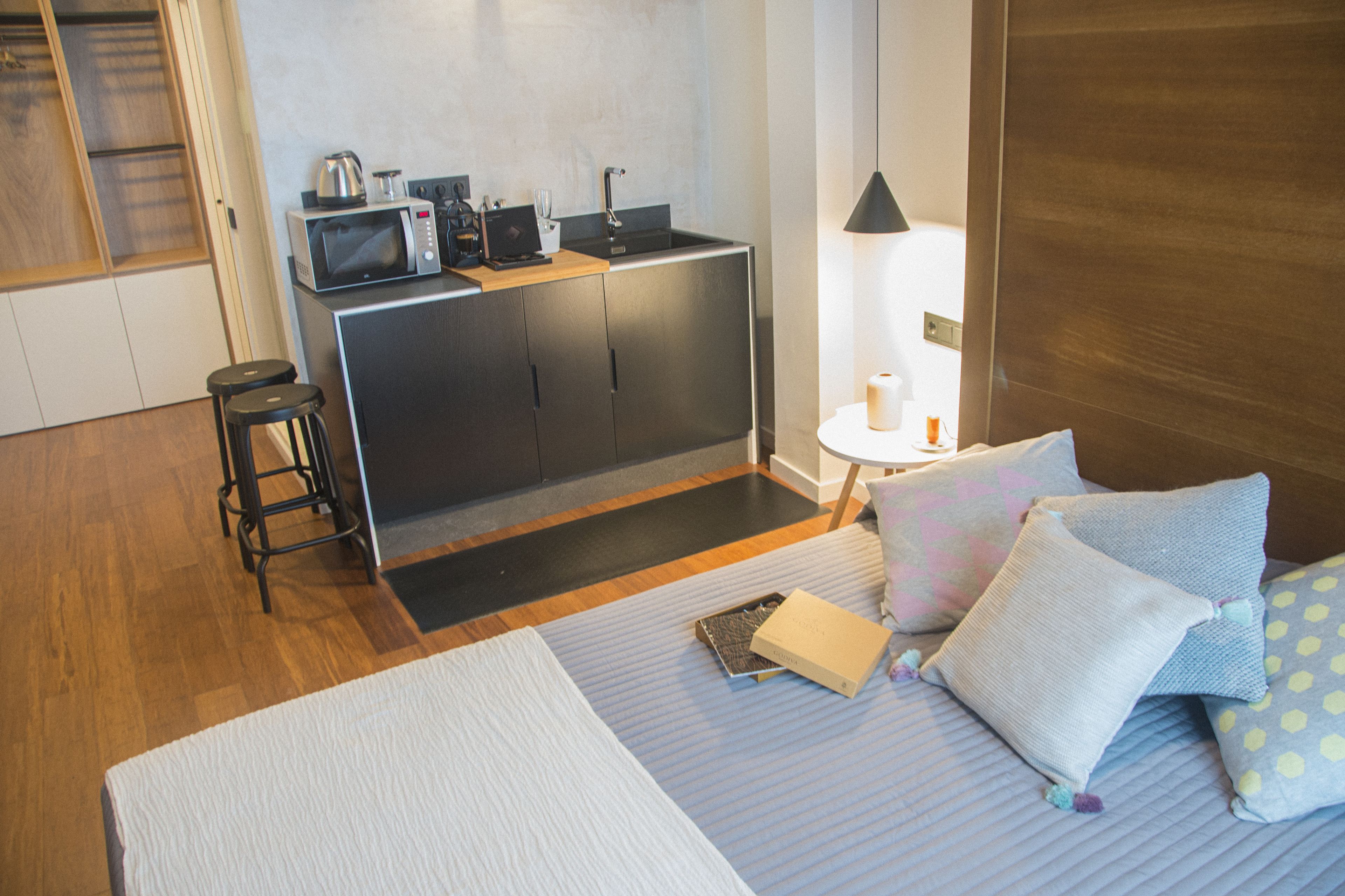 A compact hotel room with a kitchenette, a bed with pillows, a small stool, and a nightstand with a lamp.