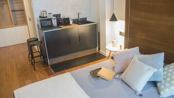 A compact hotel room with a kitchenette, a bed with pillows, a small stool, and a nightstand with a lamp.