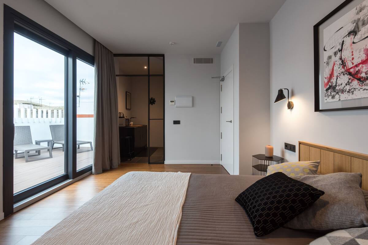 A modern hotel room with a large bed, a view of the sea through floor-to-ceiling windows, a small kitchenette, and a balcony with outdoor seating.