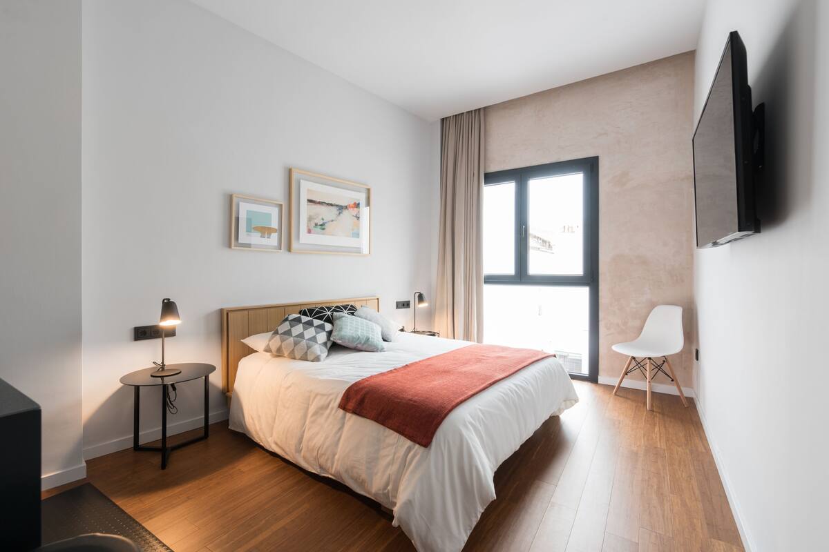 A modern bedroom with a large bed, a TV mounted on the wall, and a window with curtains.