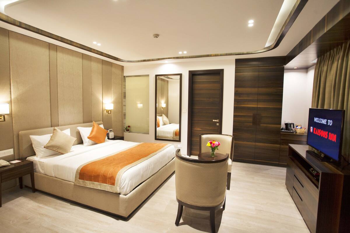 Quarto de hotel moderno com uma cama grande, mesa de cabeceira, cadeira, televisão e espelho.