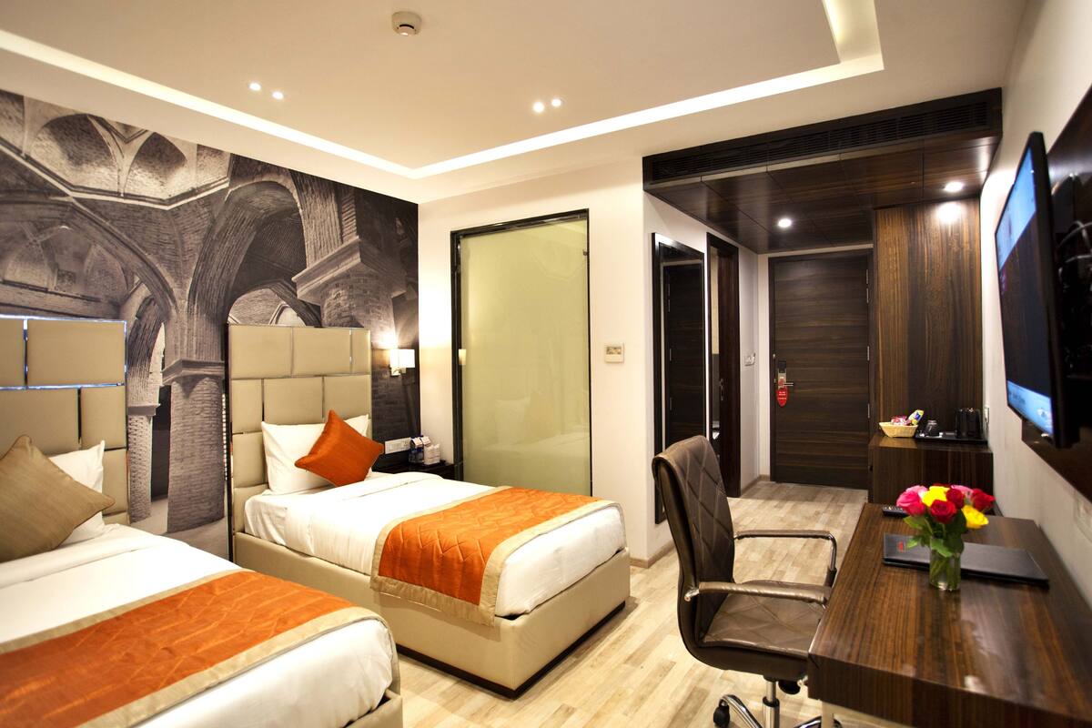 Quarto de hotel com duas camas, uma escrivaninha, uma cadeira, uma televisão e um mural na parede.