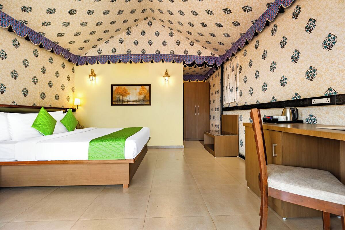 A hotel room with a bed, a desk, a chair, a wall-mounted lamp, and a painting.