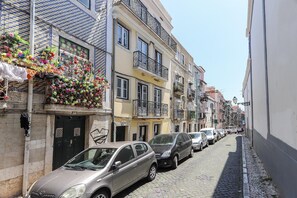 Una estrecha calle adoquinada flanqueada por edificios de varios pisos, coches aparcados y balcones decorados con flores de colores.