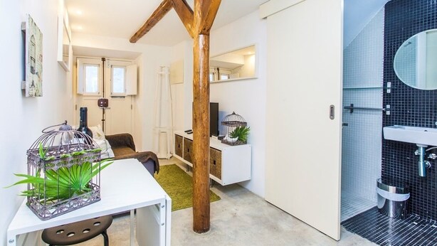 A modern interior with a white table, a wooden beam structure, a white cabinet, and a bathroom with black tiles and a mirror. - Bairro Alto Green by Homing (Lisbon)