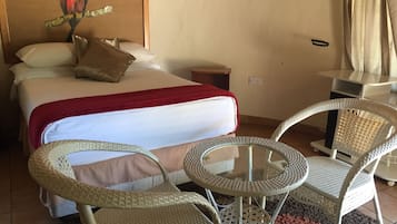A hotel room with a bed, two wicker chairs, a small round table, a red throw blanket, and a painting of a bird on the wall.