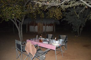 An outdoor dining setup with a table set for two, surrounded by chairs, under a tree. - Masama Lodge and Campsite (Serowe)