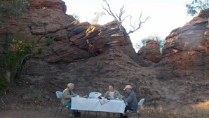 Un grupo de personas cenando en una mesa al aire libre con un fuego en frente, frente a un paisaje rocoso.