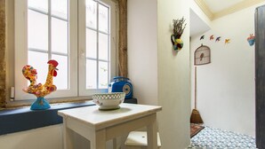 A small room with a window, a white table, a colorful rooster decoration, a blue radio, and a birdcage on the wall.