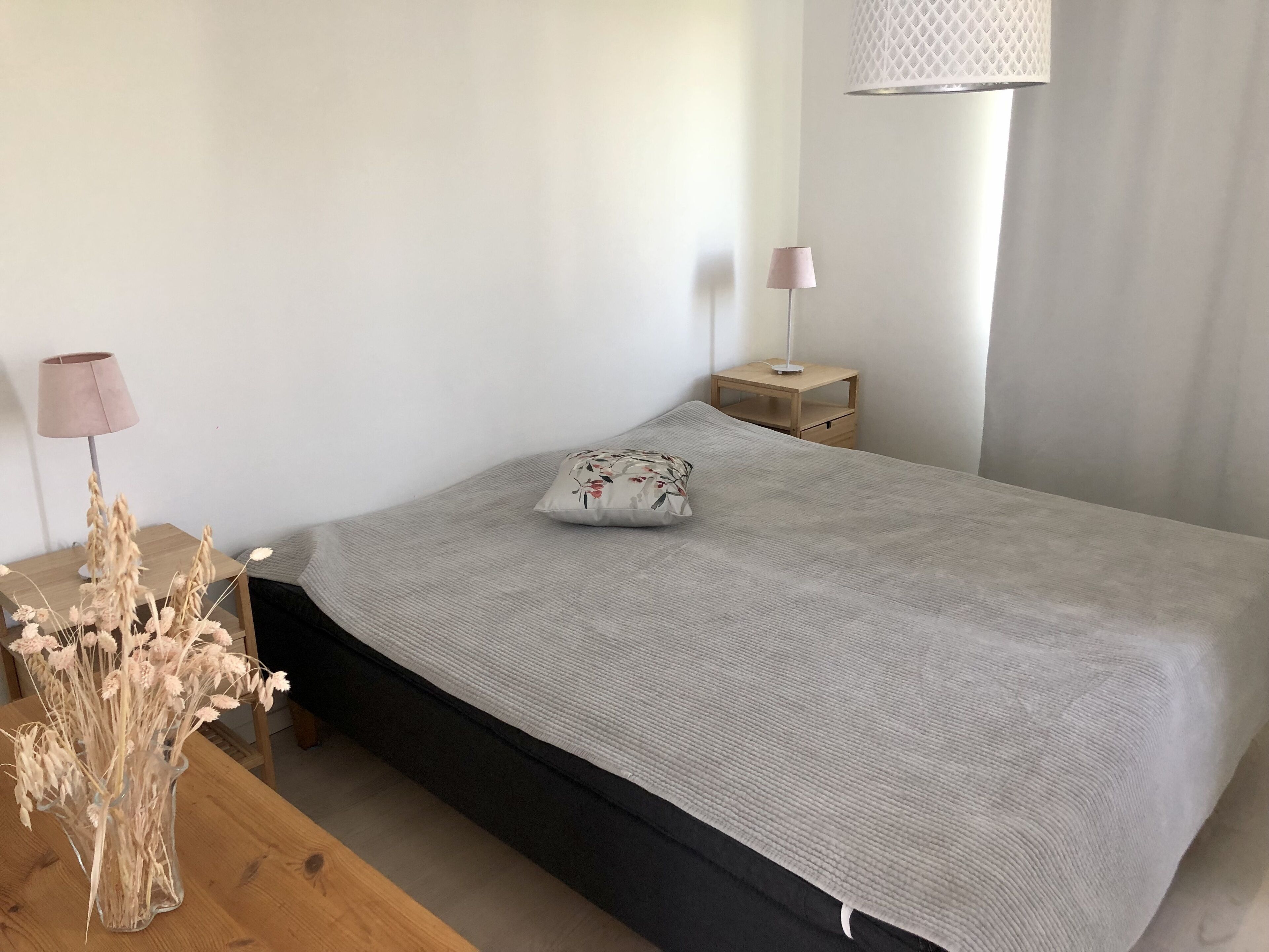 A neatly made bed with a grey blanket, a floral pillow, and a bedside lamp on a wooden nightstand.
