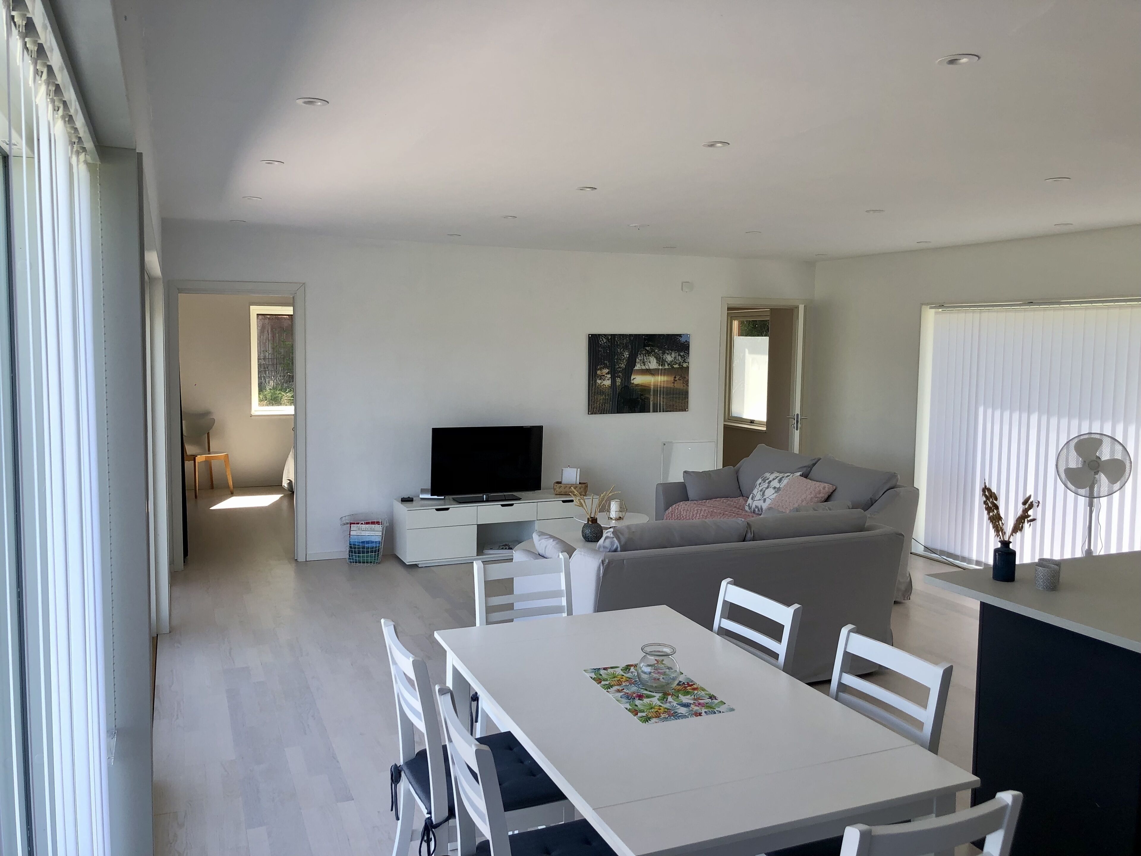 Ett modernt vardagsrum med en vit soffa, en platt-TV och en matplats med ett vitt bord och stolar.