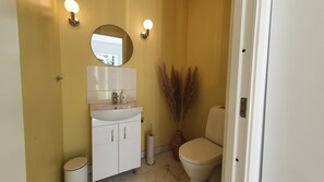 Un baño con lavamanos, espejo y retrete blancos. Hay dos luces empotradas en la pared y un pequeño cubo de basura.