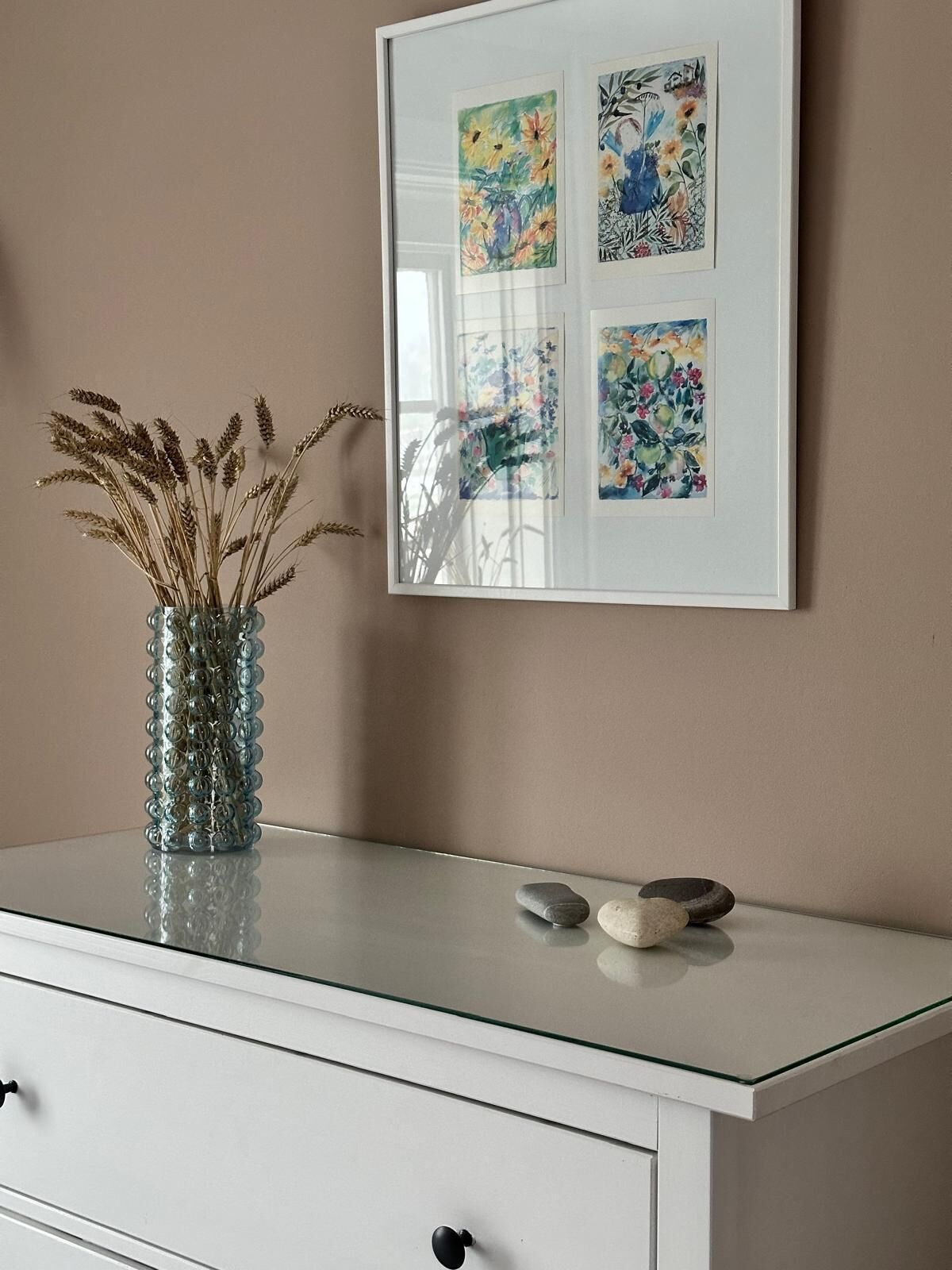 A white dresser with a glass vase of dried wheat stalks, two smooth stones, and a framed floral artwork on the wall above.