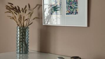 A white dresser with a glass vase of dried wheat stalks, two smooth stones, and a framed floral artwork on the wall above.