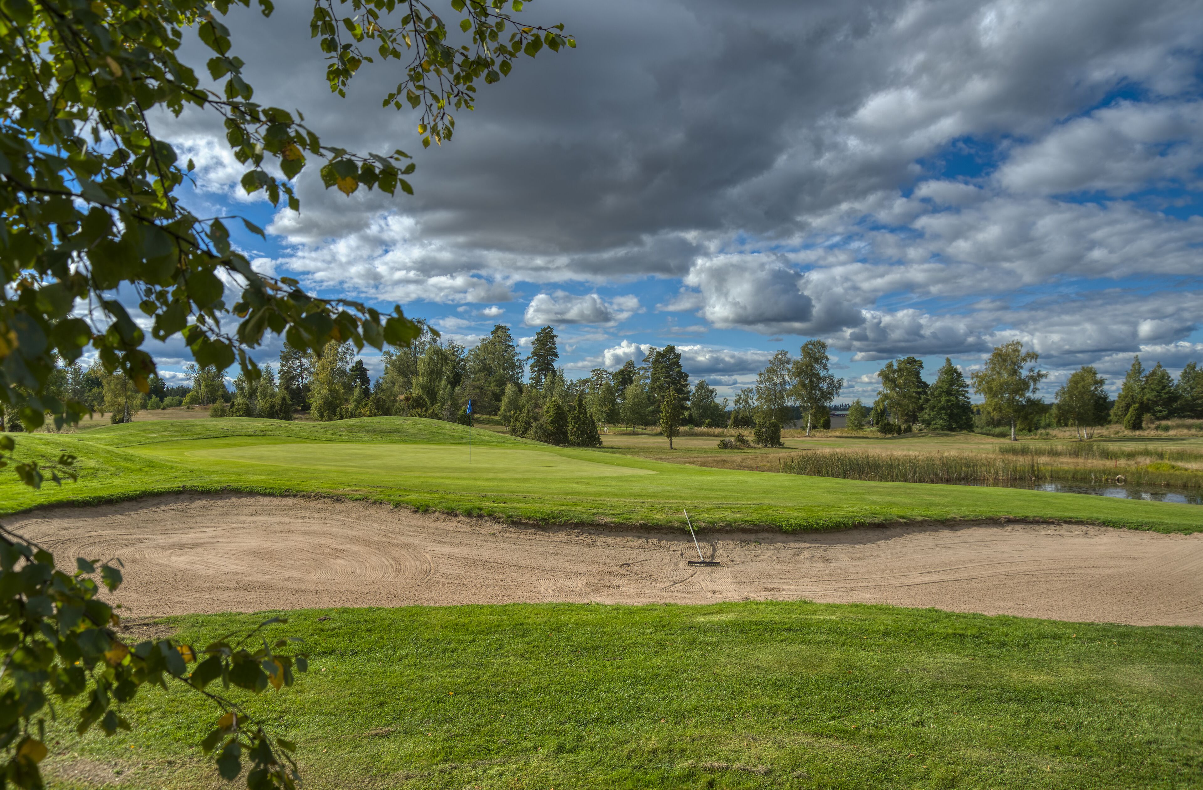 A golf course with a water hazard, trees, and a partly cloudy sky.