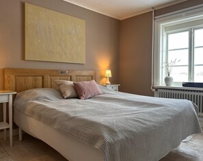 Un dormitorio con una cama grande, una mesita de noche con lámpara, una ventana con vistas y un cuadro en la pared.
