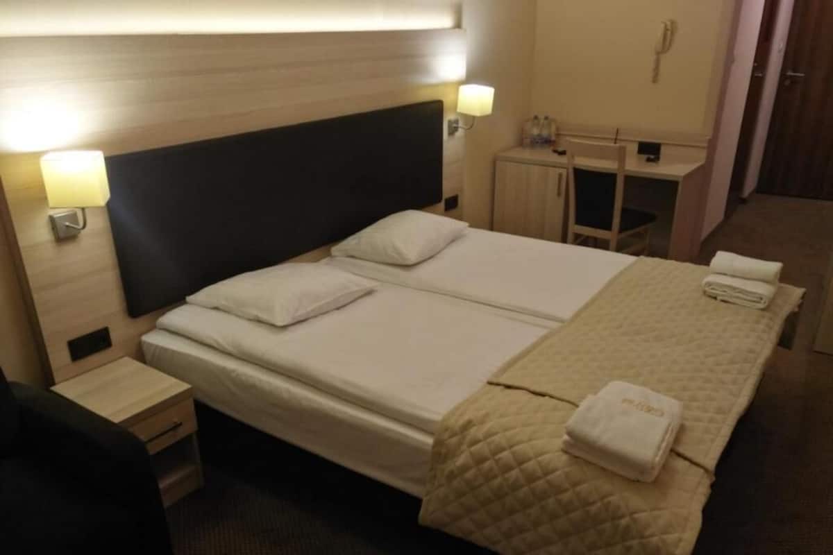A double bed with a quilted blanket, a bedside table, a nightstand, a desk, and a wall-mounted telephone.