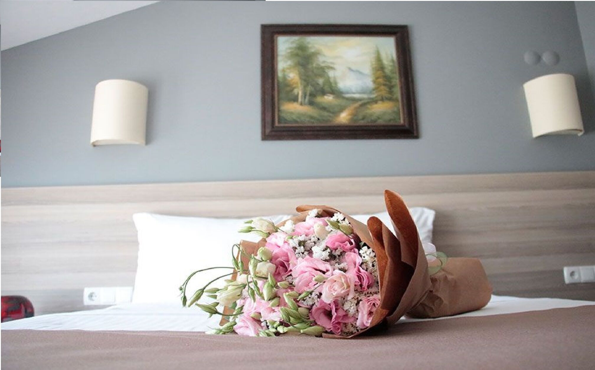 Een hotelkamer met een bed, een bloemenboeket, een schilderij aan de muur en twee wandlampen.