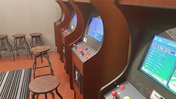 A row of arcade gaming machines with multiple joysticks and screens, wooden stools, and a patterned rug.