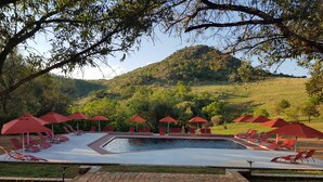 Zona de piscina con sombrillas rojas y sillas de descanso, rodeada de vegetación y colinas.