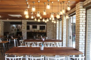 Un restaurante con mesas y sillas de madera, lámparas colgantes, paredes de ladrillo y ventanas amplias.