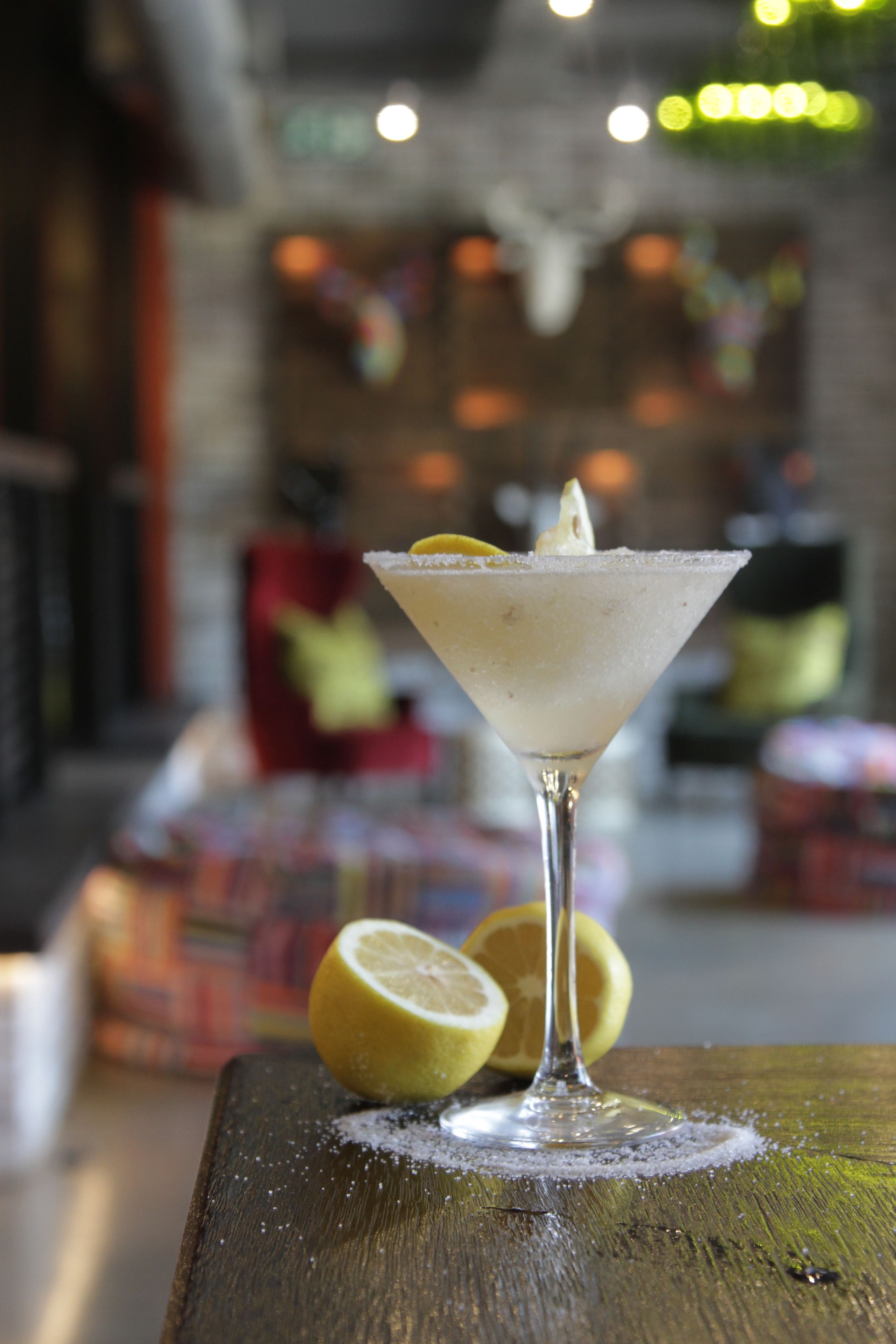 Ein Martini-Glas mit einer Zitronenscheibe auf einem Holztisch, im Hintergrund verschwommen ein Barbereich.