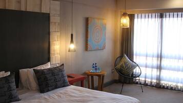 Habitación de hotel con cama, una mesita con botellas de agua, una silla y un cuadro en la pared.