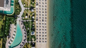 Vista aérea de un complejo turístico en la playa con filas de tumbonas y sombrillas, una piscina y un edificio con techo curvo.