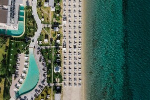 Vista aérea de un complejo turístico en la playa con filas de tumbonas y sombrillas, una piscina y un edificio con techo curvo.