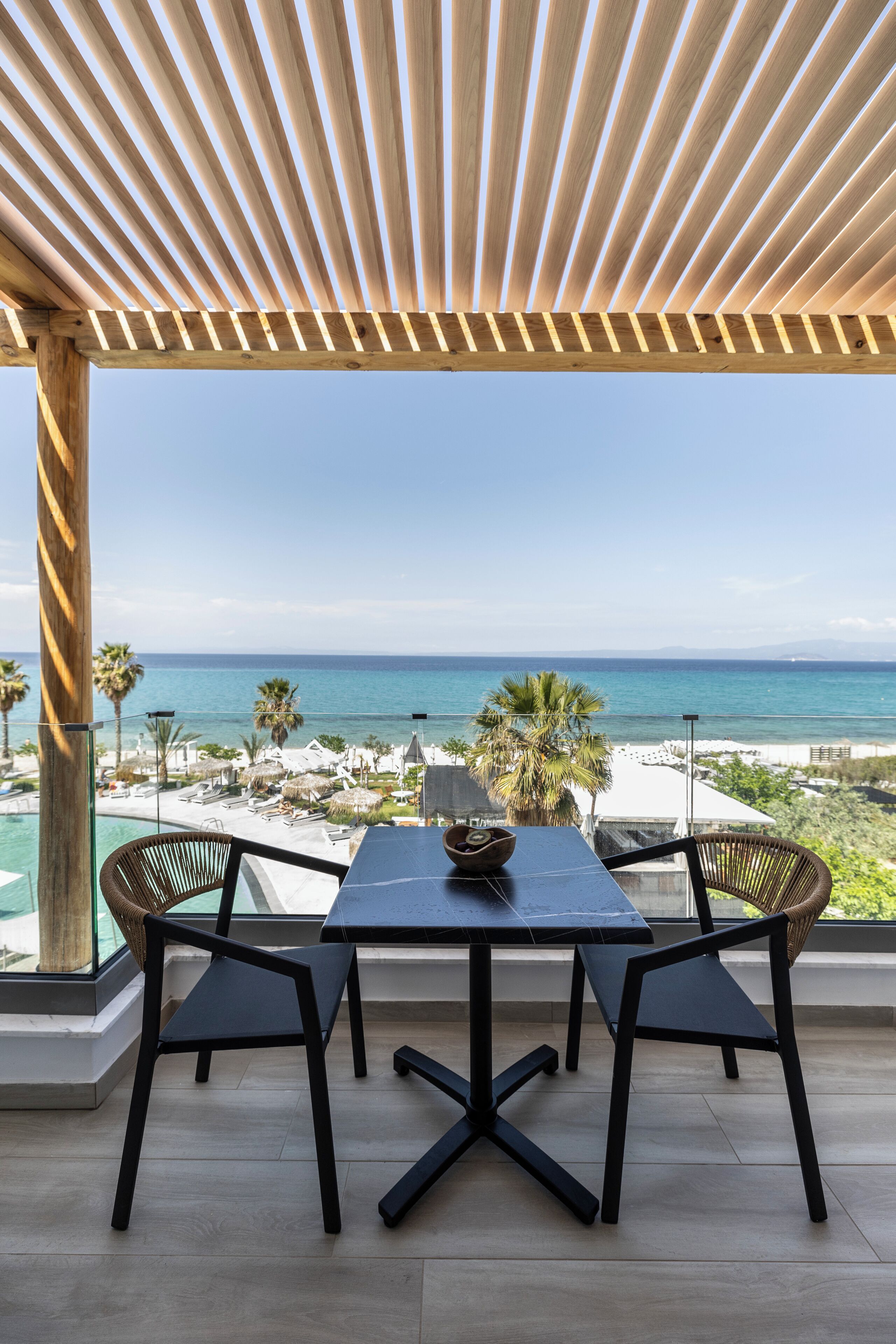 Una terraza con pérgola de madera, una mesa, dos sillas y vista al océano y la playa.