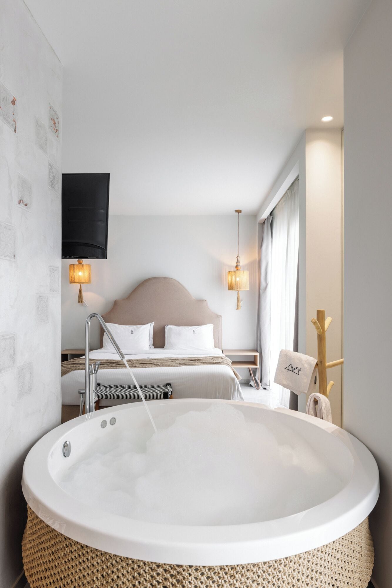 Habitación de hotel moderna con una bañera grande, una cama con cabecera de madera, dos lámparas de pared y un televisor de pantalla plana.