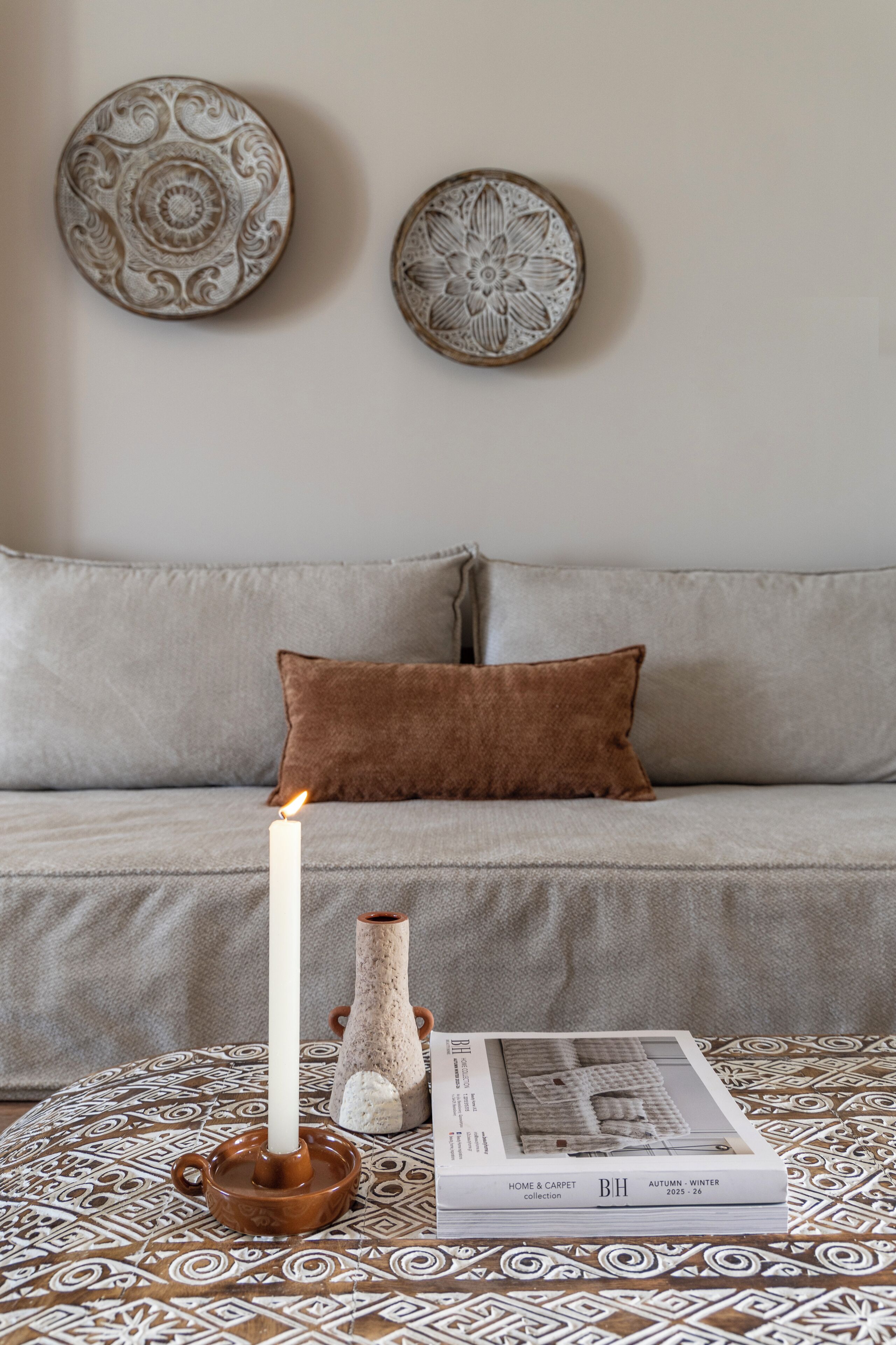 A living room with a sofa, a brown cushion, a table with a candle, a vase, and a magazine.