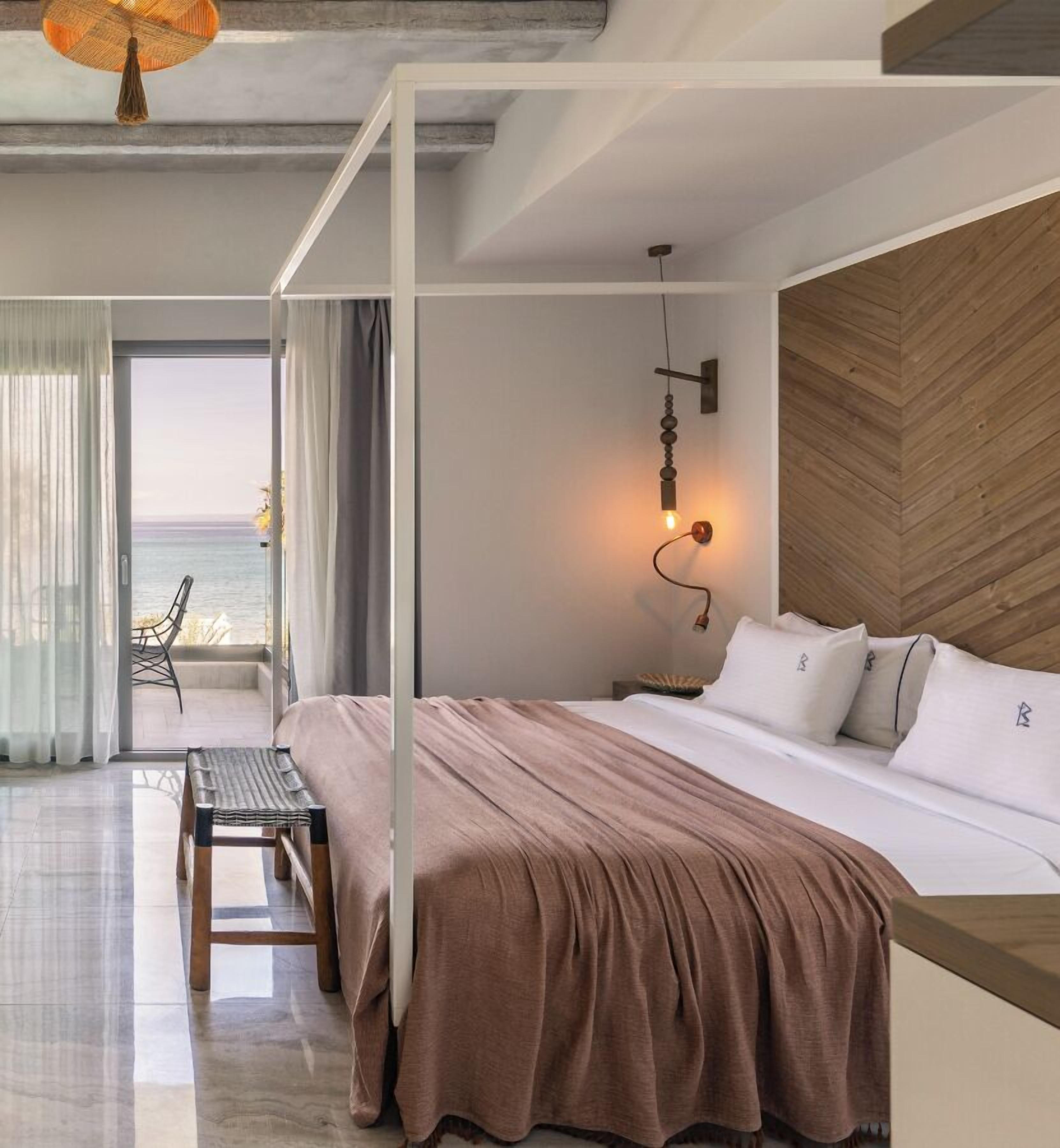 A modern bedroom with a large bed, a wooden headboard, a bedside table, and a view of the sea through a glass door.