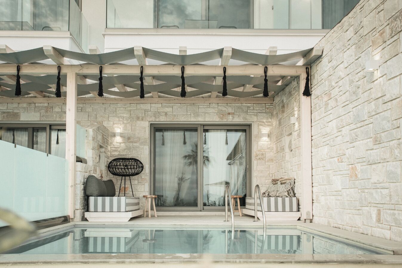 A modern outdoor pool area with a stone wall, a covered seating area, and a glass door leading to the pool.