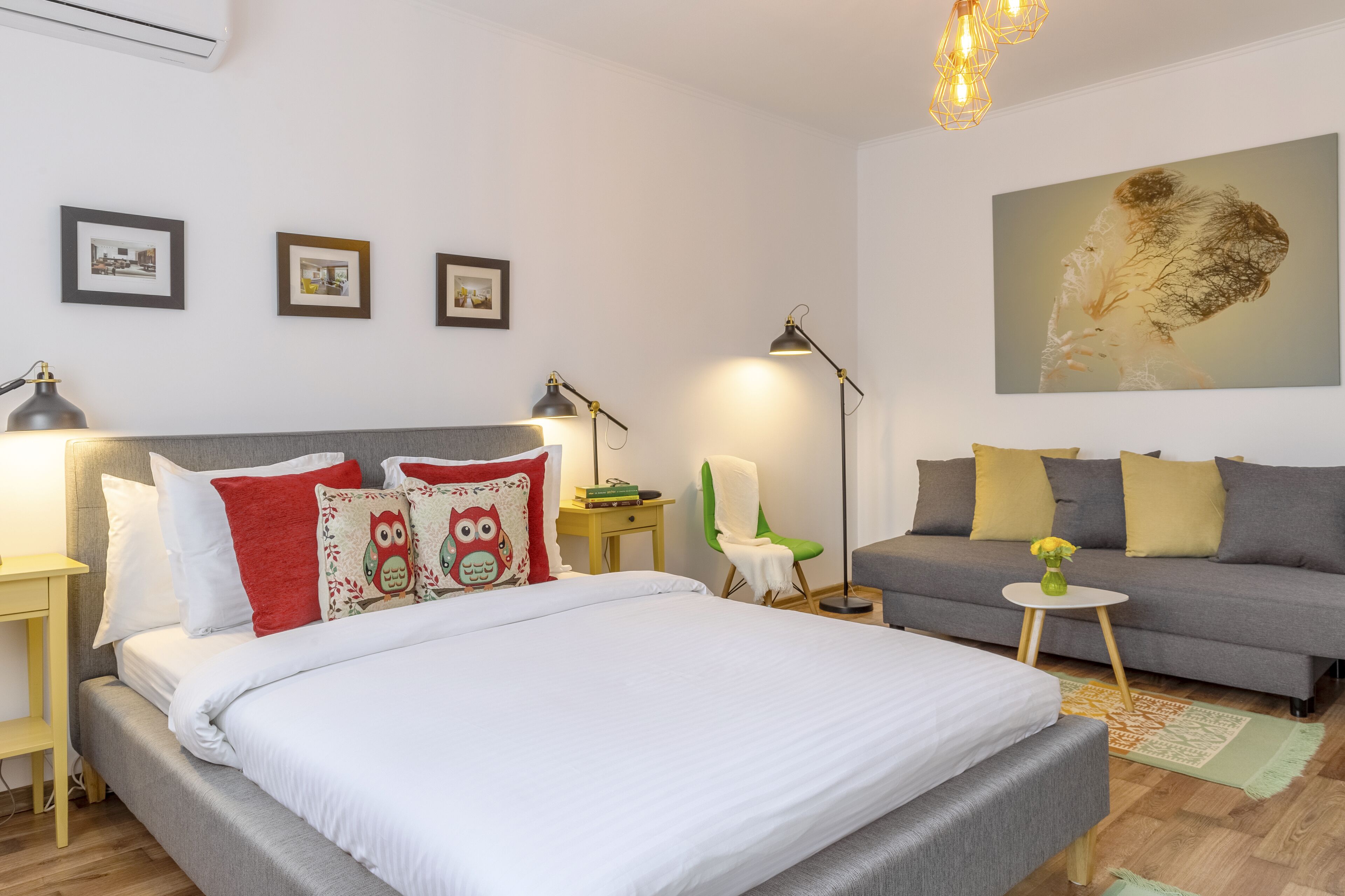 A well-lit bedroom with a bed, a sofa, a chair, and a small table. There are framed pictures on the wall and a large painting above the sofa.
