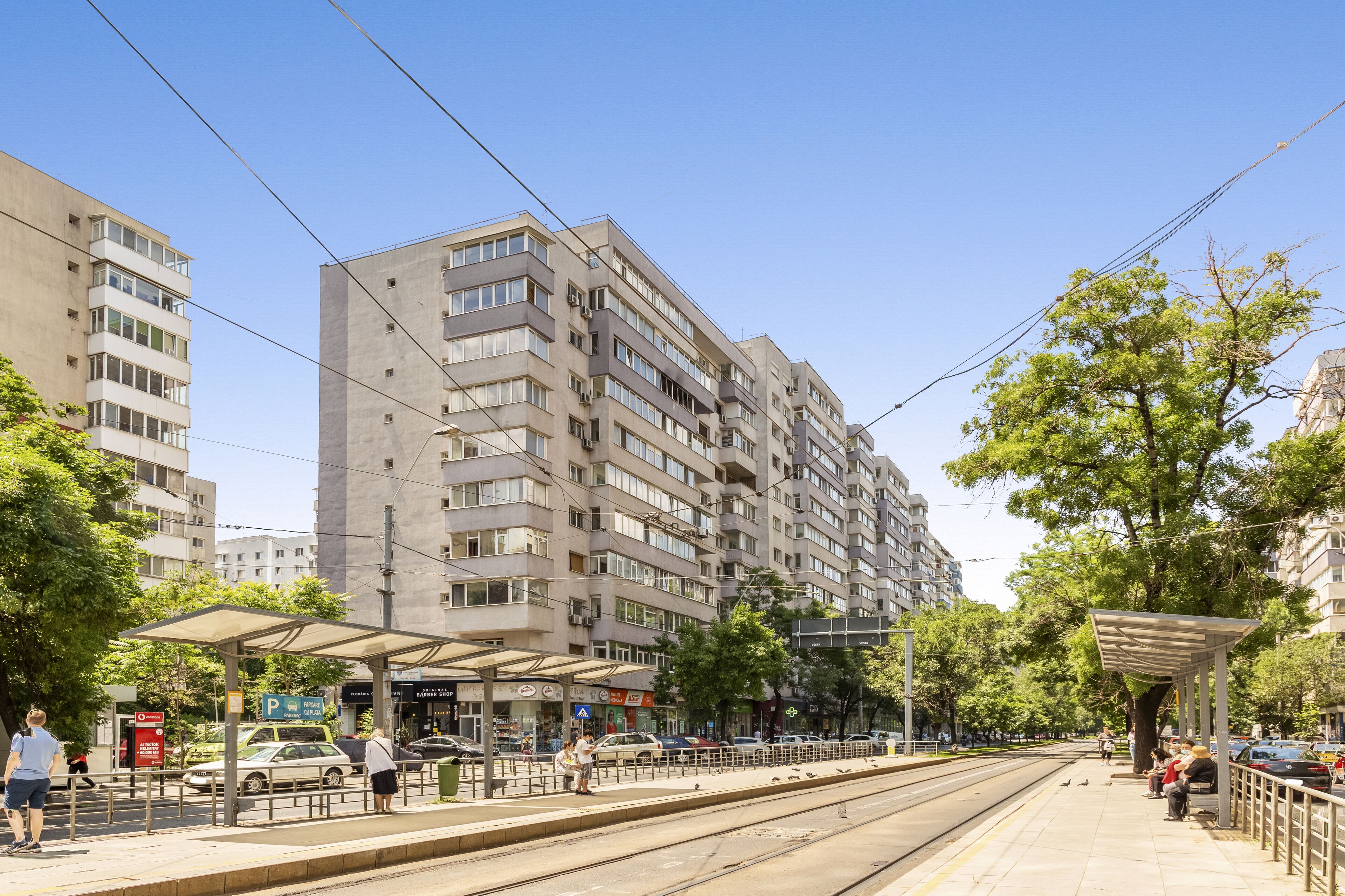 A multi-story residential building with balconies, a tram line, and a pedestrian walkway with a covered shelter.