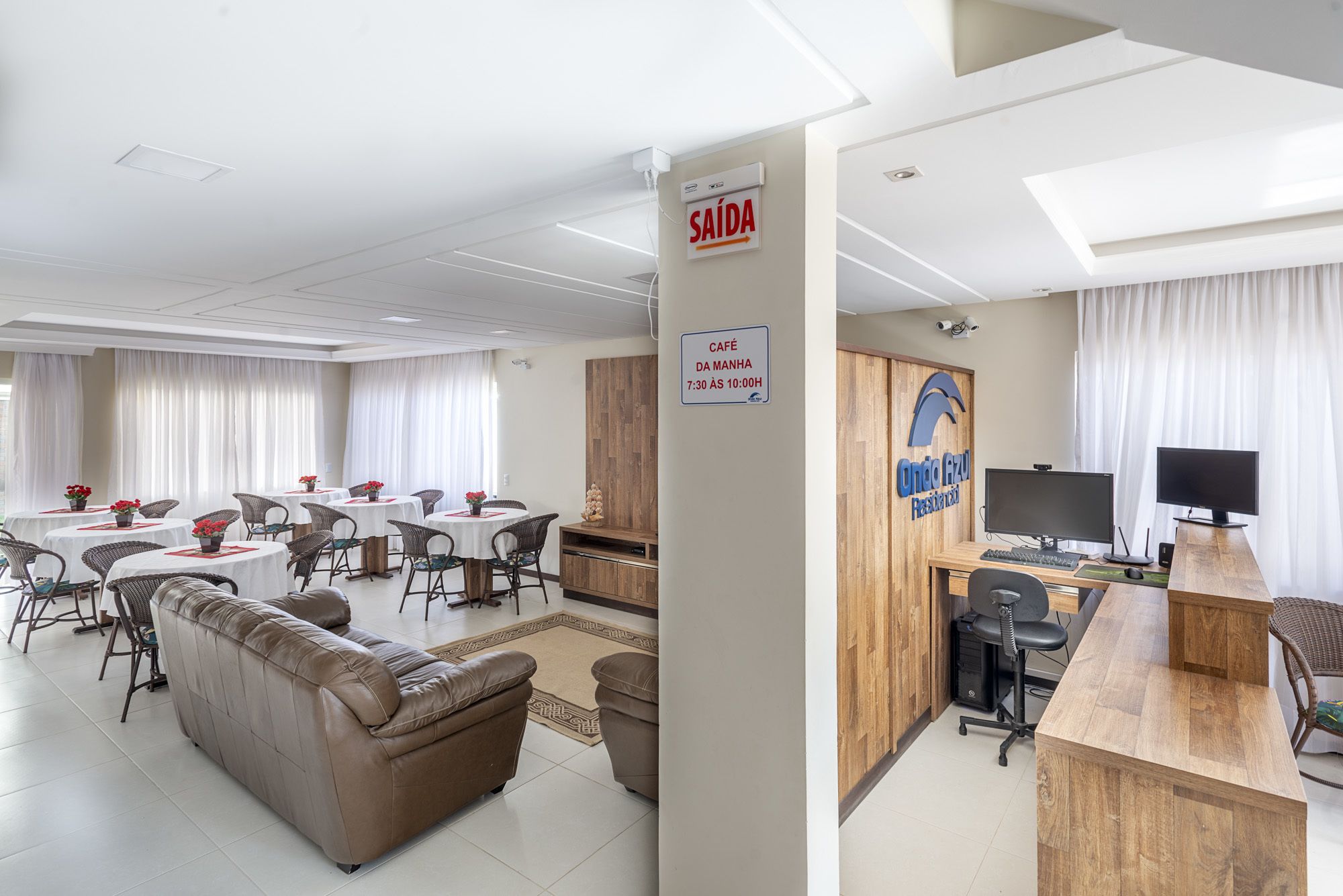 Un hall d’hôtel comprenant un comptoir d’accueil, un espace repas avec des tables et des chaises rondes, un canapé et un panneau indiquant les heures d’ouverture du café.