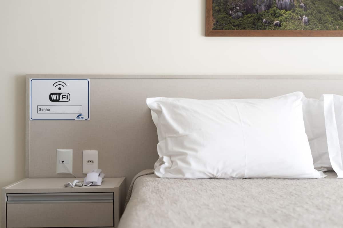 A hotel room with a bed, a nightstand with a phone and a Wi-Fi sign, and a painting on the wall.