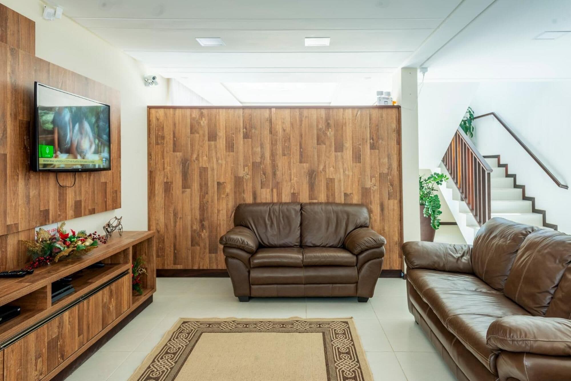 Una sala de estar con pared de madera, un sofá de cuero marrón, un televisor en la pared y una escalera al fondo.