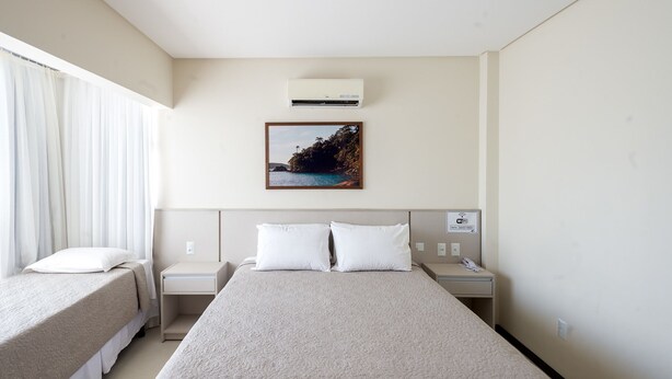 A hotel room with a large bed, two bedside tables, a nightstand, a wall-mounted air conditioner, and a framed picture of a coastal landscape.