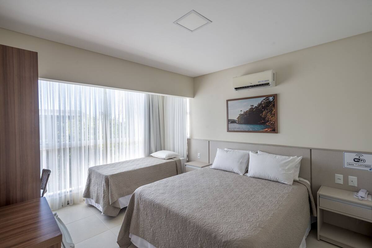 A hotel room with two beds, a large window with sheer curtains, an air conditioning unit, a Wi-Fi sign, and a framed picture on the wall.