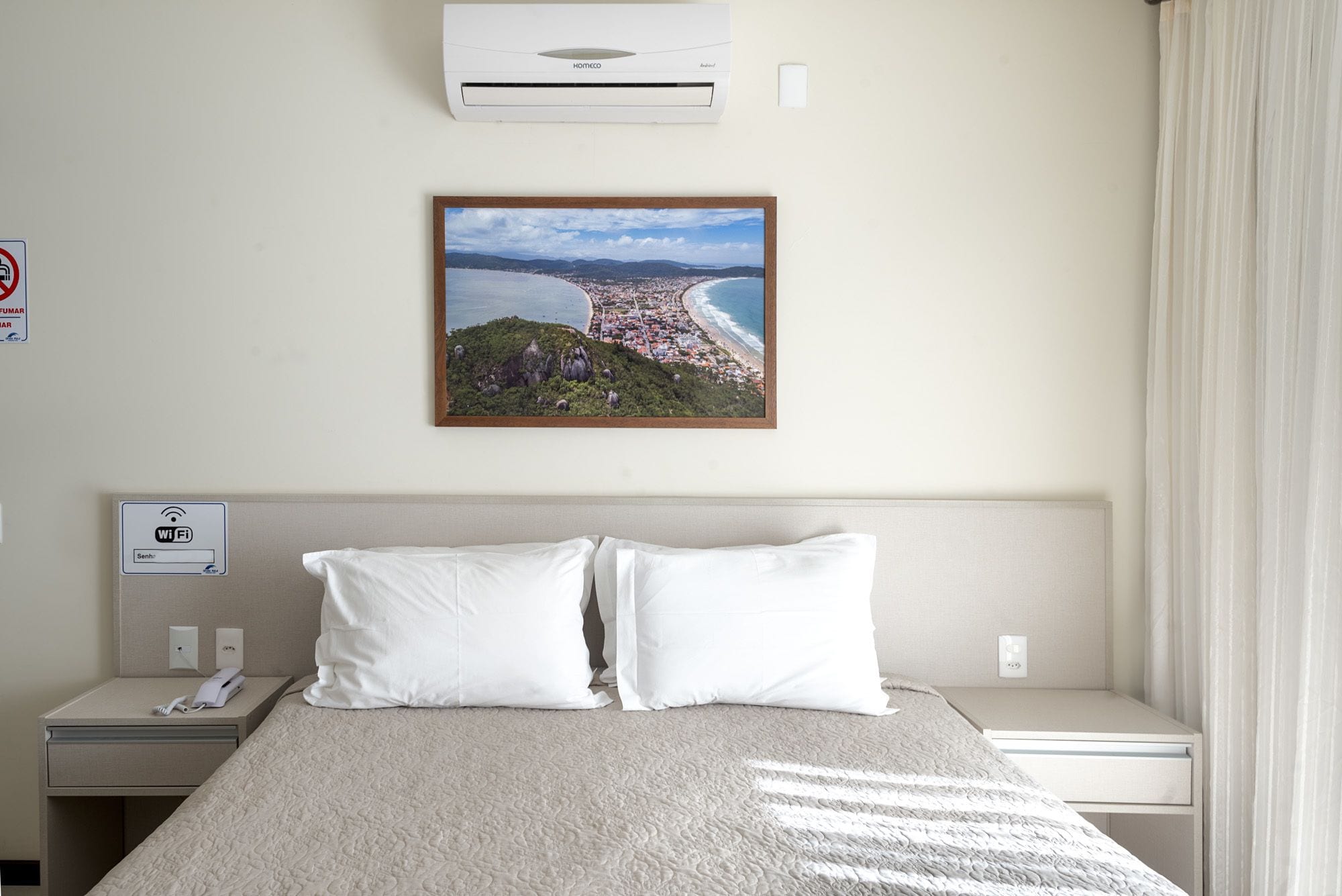 Et hotelværelse med en seng, et natbord med en telefon, en aircondition og et billede i ramme af en kystby.