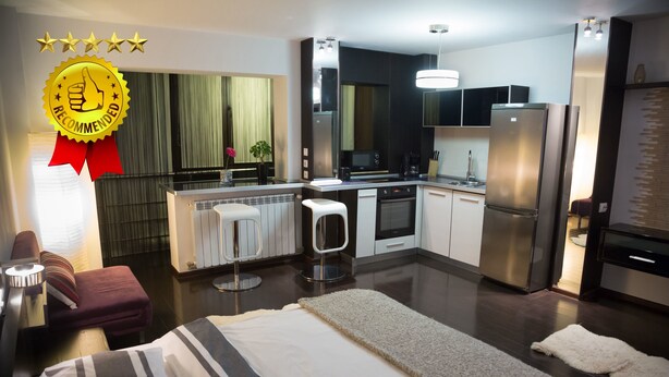 Cocina moderna con refrigerador, estufa y microondas. Hay un pequeño comedor con una mesa y sillas. La habitación tiene una cama con ropa de cama a rayas y un amplio ventanal con cortinas.