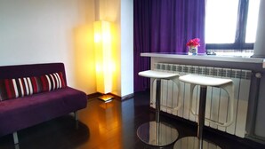 Una sala de estar moderna con un sofá morado, una lámpara de pie y una zona de cocina con ventana y cortinas moradas.