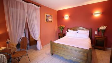 A bedroom with a wooden bed, white curtains, a small table with chairs, and a wall-mounted light fixture.