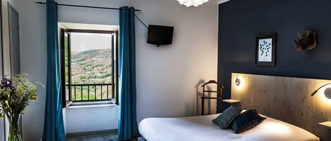 A bedroom with a large bed, a view of a hilly landscape, a TV on the wall, and a modern light fixture.
