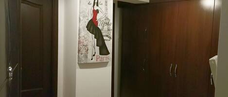 Un couloir avec une armoire en bois sombre, une porte et une affiche murale représentant une femme en robe, sur laquelle on peut lire les mots « Salon de la Mode Paris ».
