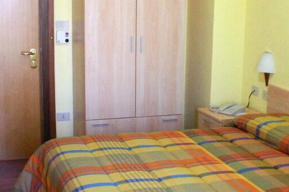 Habitación de hotel con cama, armario, mesita de noche con lámpara y teléfono en la pared.