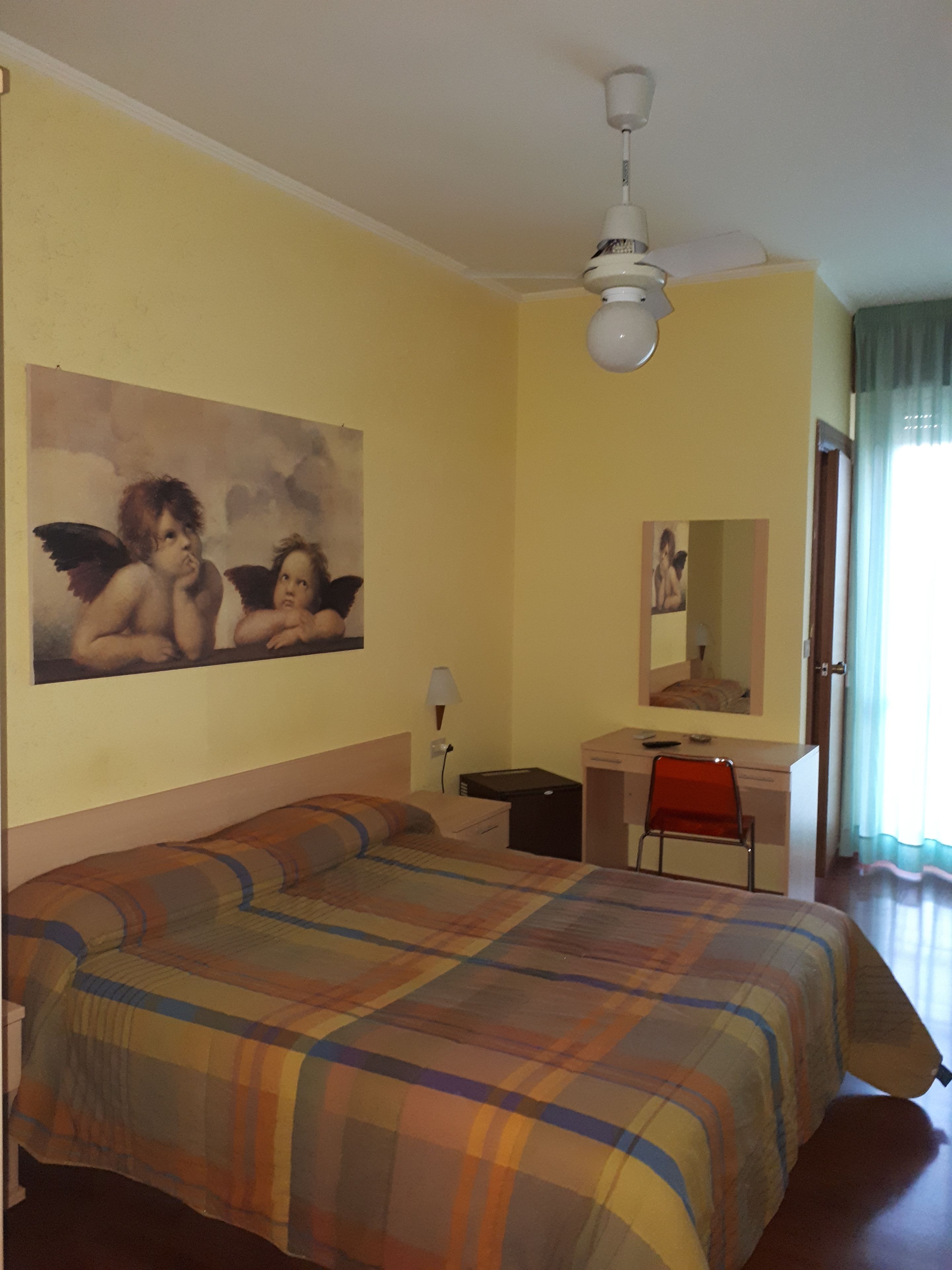 Una camera d'albergo con un letto, una scrivania, una sedia, una ventola a soffitto, una lampada a parete e un dipinto di cherubini.